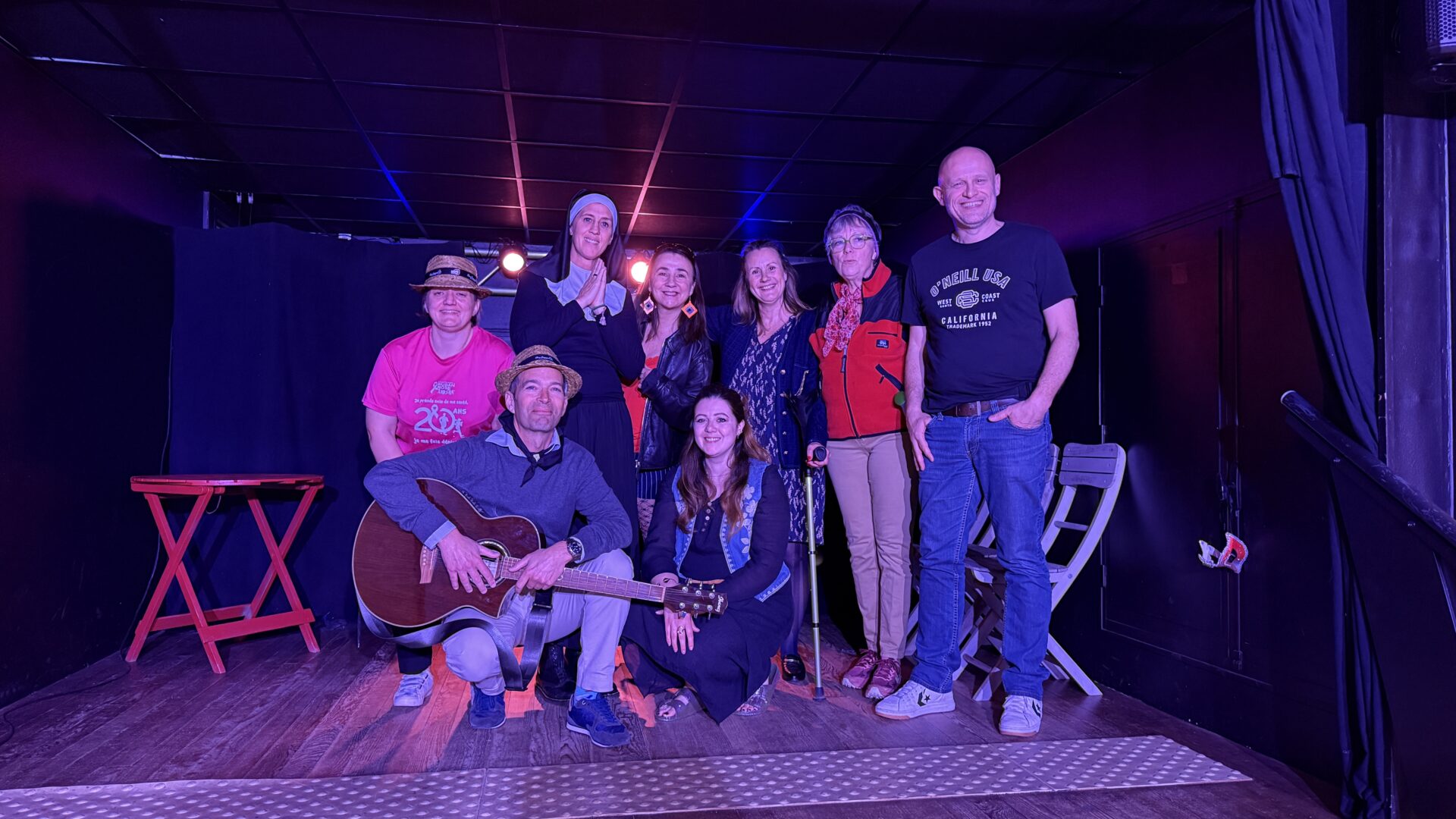 Un groupe de personne dont un homme tenant une guitare au premier plan prend la pose sur scène, éclairage rose.