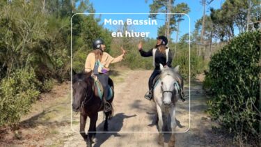 Deux jeunes femmes l'une en veste beige et l'autre noir sont sur des chevaux marron et blanc dans un chemin sableux en forêt