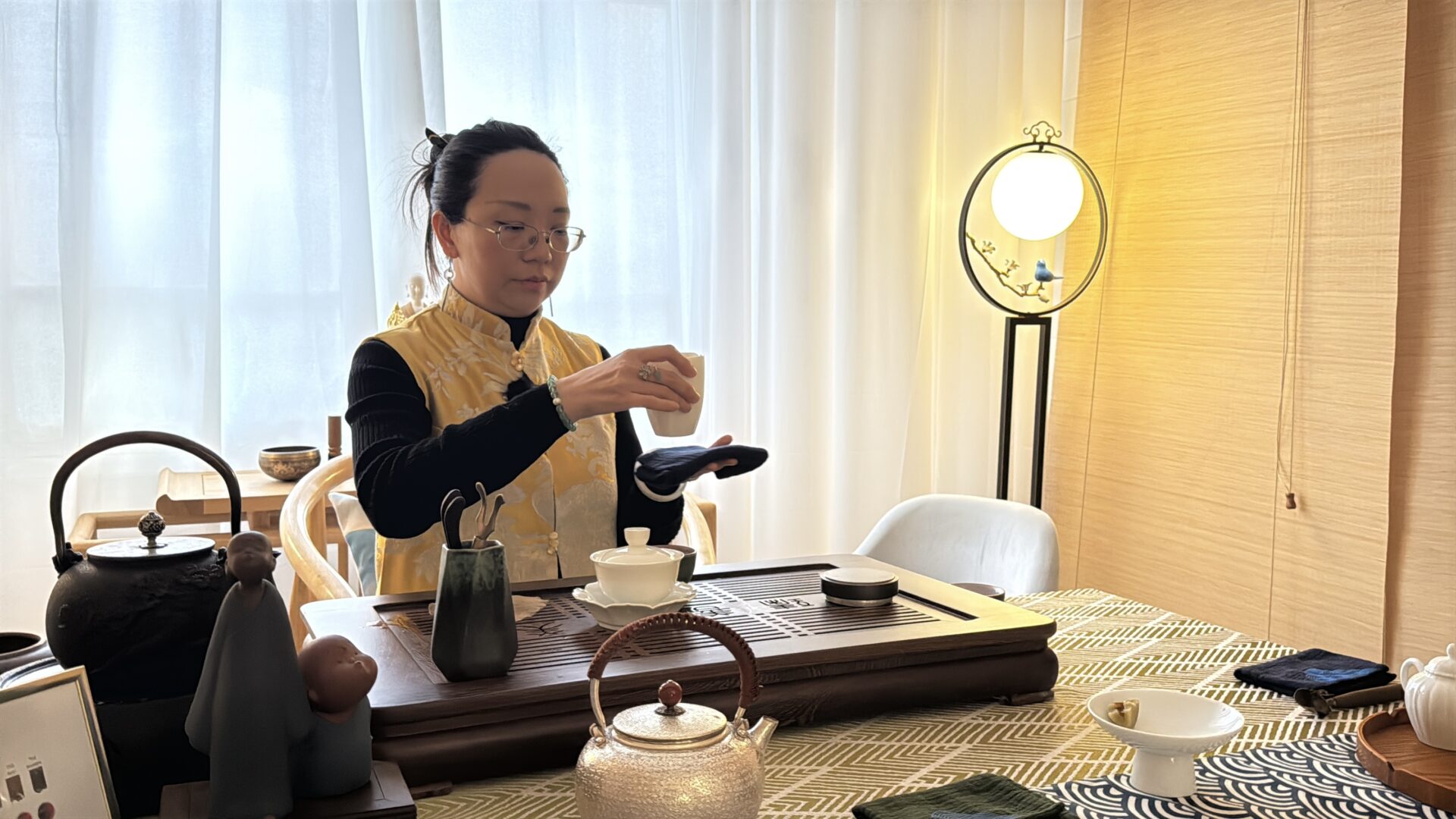 Une femme prépare du thé sur un plateau avec théière et tasses, dans une pièce lumineuse et calme.