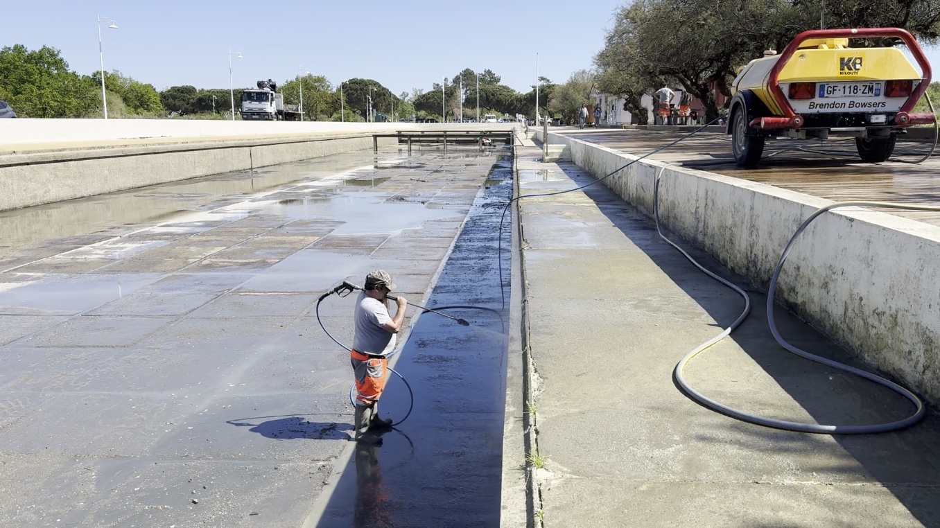 Un homme en pantalon orange manie un puissant jet d'eau pour nettyer un grand bassin, type piscine en béton de plein air