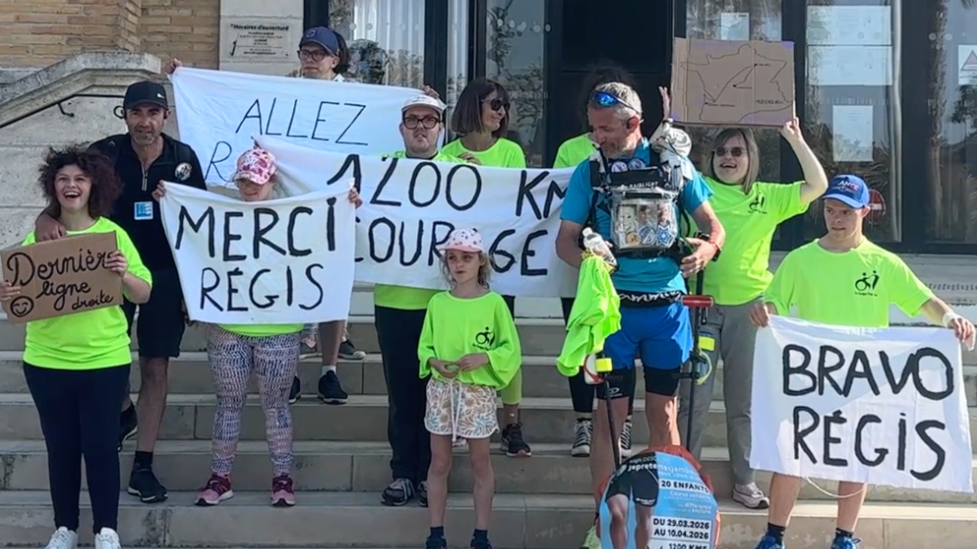 Un groupe d’adultes et d’enfants en t-shirts verts encourage un coureur équipé, en tenant des pancartes « Bravo Régis », « Merci Régis » et « 1200 km »