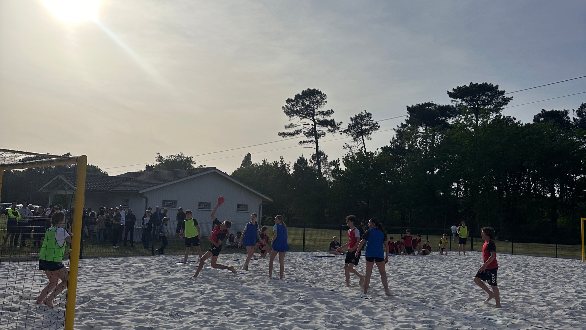 Un groupe de jeunes joue au beach handball sur un terrain de sable en plein air. Un joueur en rouge saute pour tirer, pendant que d’autres défendent et que le gardien surveille le but.