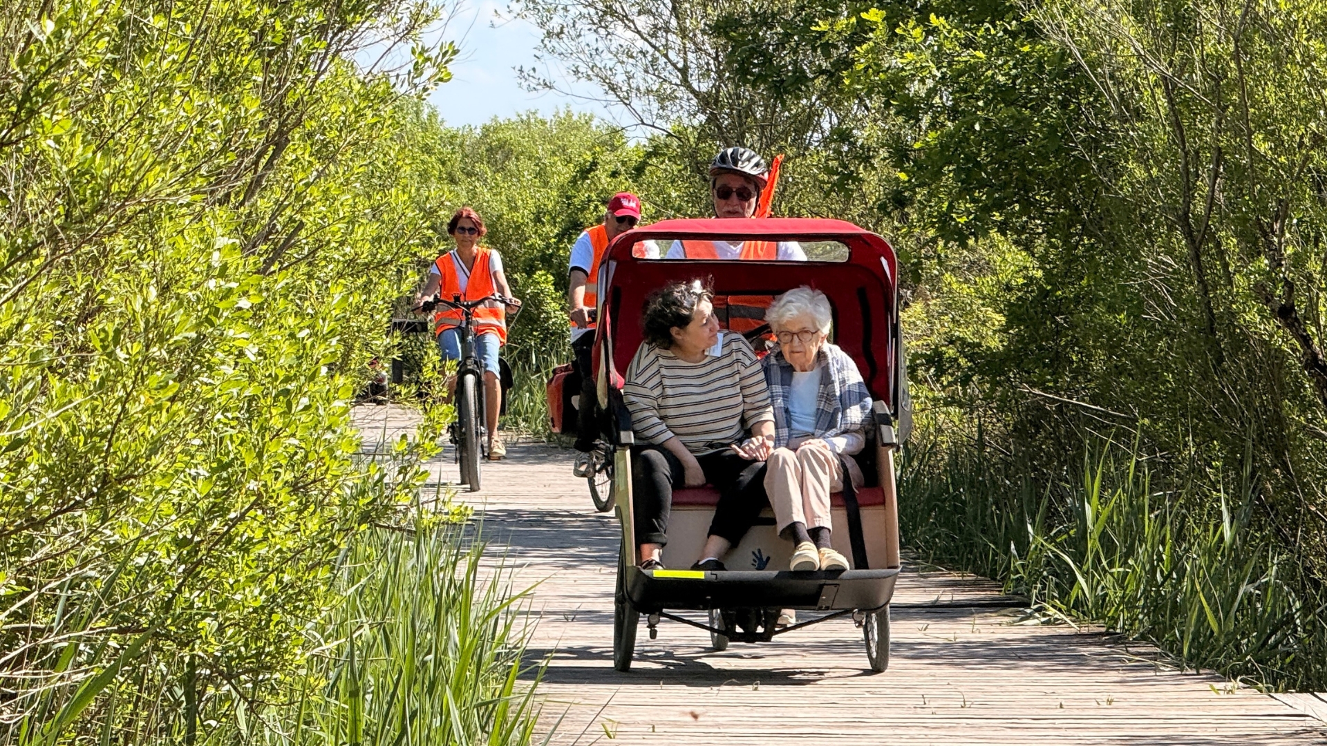 Sur un chemin au milieu de la nature, une senior et son accompagnante dans un triporteur rouge conduit par un homme, en arrière plan des personnes sur des vélos avec des gilets orange