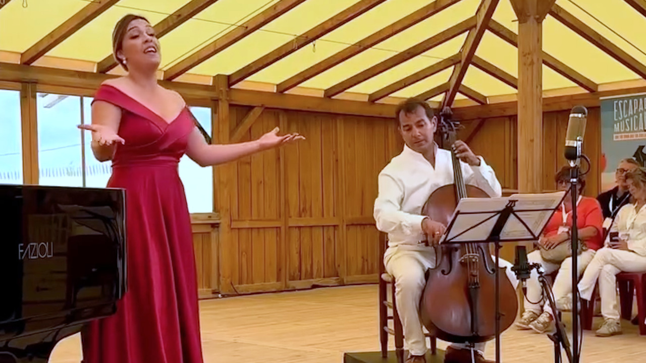 Une chanteuse d'opera en robe rouge et un musicien en chemise blancge dans une piece au mur de bois et au toit en toile.
