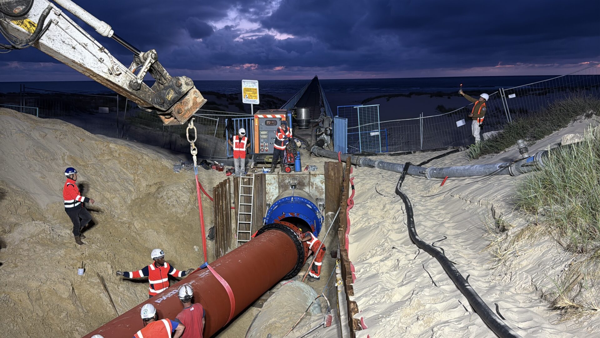 Un chantier devant la plage sur une dune de sable. On peut voir de gros tuyaux rouge enfoncés dans le sable, une grue et des hommes en orange qui s'affairent