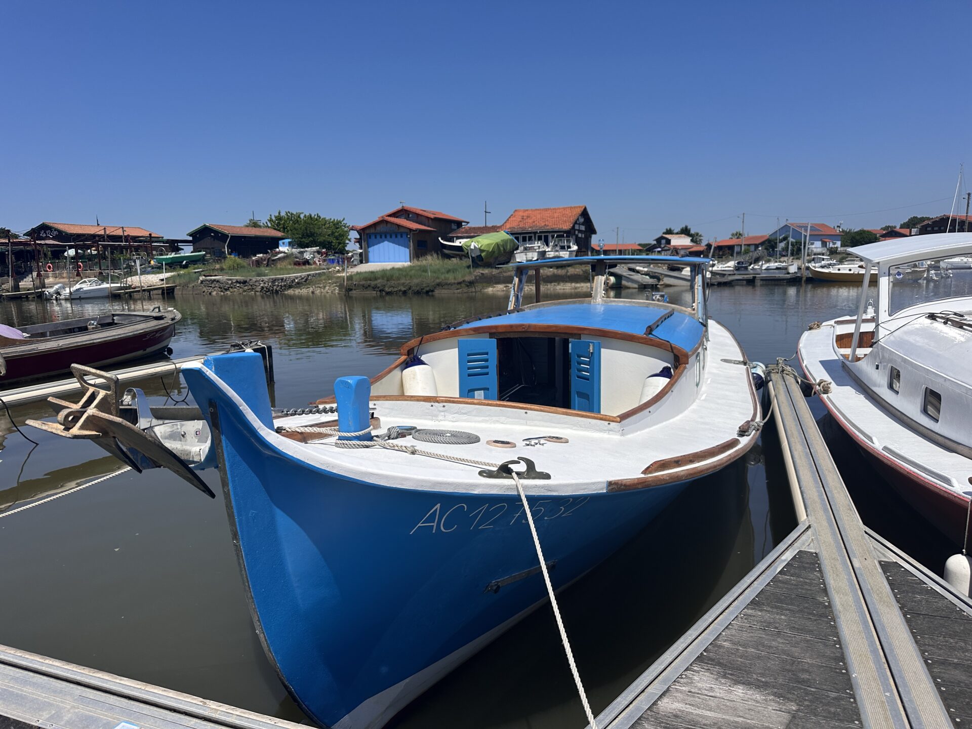 Une pinasse bleue est amarrée dans le port de La Teste-de-Buch, entourée de bateaux et de cabanes ostréicoles sous un grand ciel bleu ensoleillé.