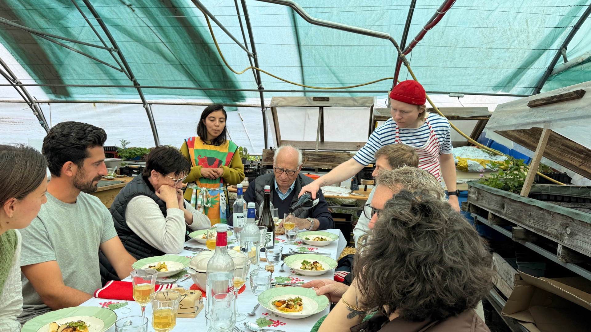 Dans la serre d'une pépinière : une table et des personnes assises sur des bottes de paille, deux personnes sont debout et les servent