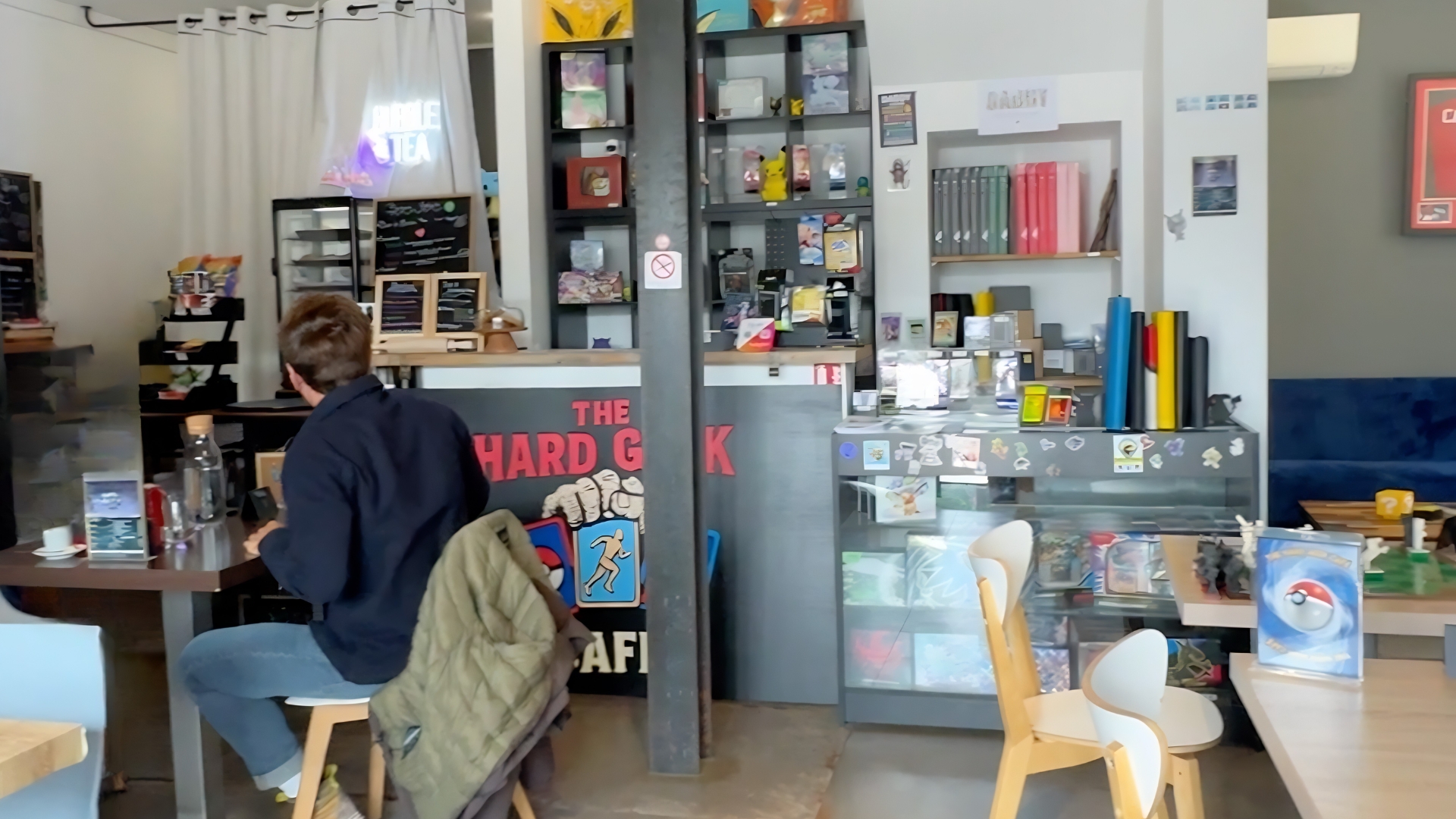 Un café à thème geek, décoré d'objets et de figurines liés à l’univers Pokémon et aux jeux vidéo, avec des étagères pleines de cartes à collectionner, de jeux et de classeurs. Un homme assis à une table regarde vers le comptoir surmonté d'une enseigne lumineuse « Bubble Tea ».