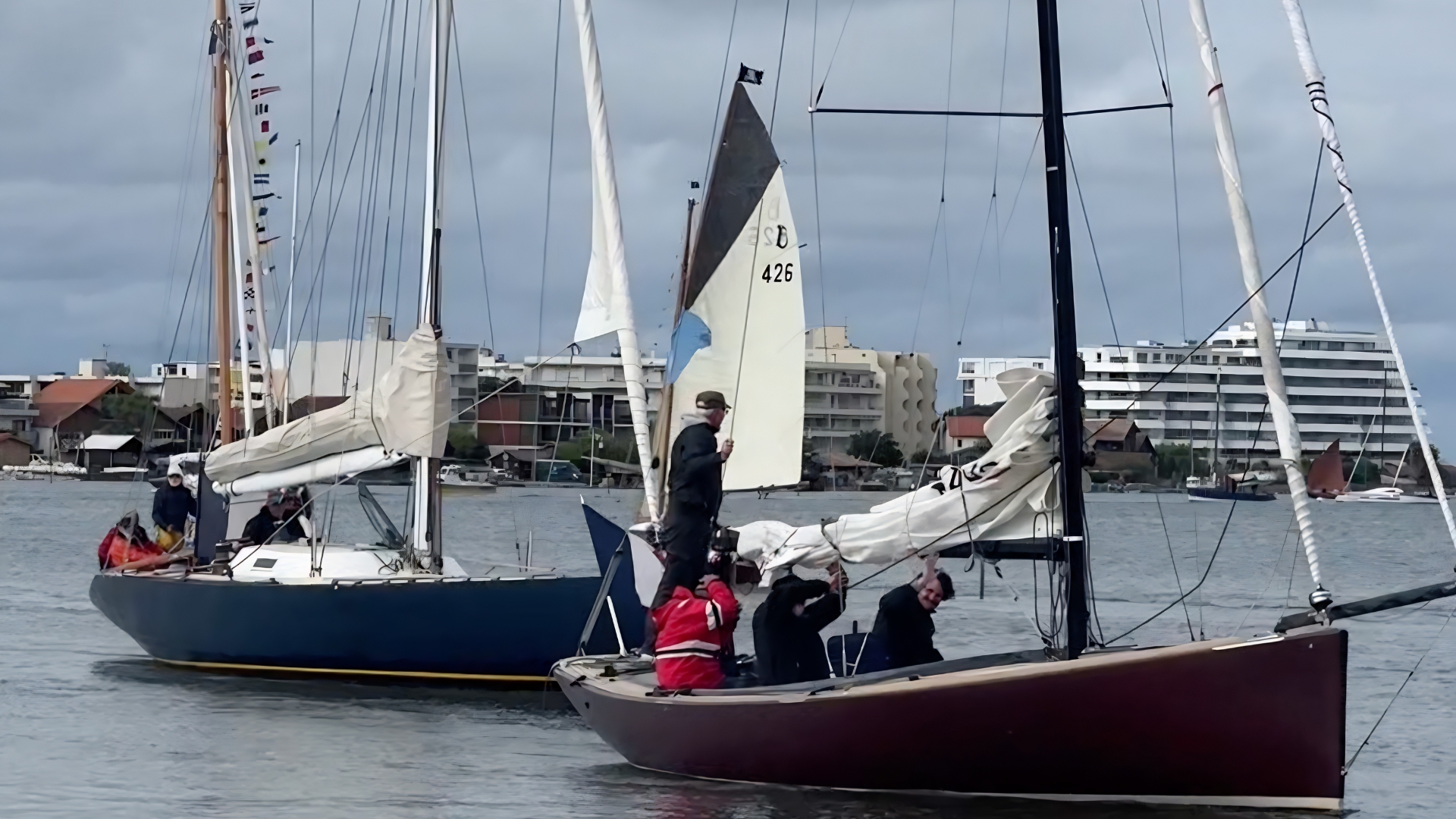 Deux voiliers classiques naviguent côte à côte dans le port de La-Teste-de-Buch, chacun avec plusieurs personnes à bord préparant les voiles. En arrière-plan, des bâtiments résidentiels modernes et d'autres bateaux sont visibles sous un ciel nuageux.