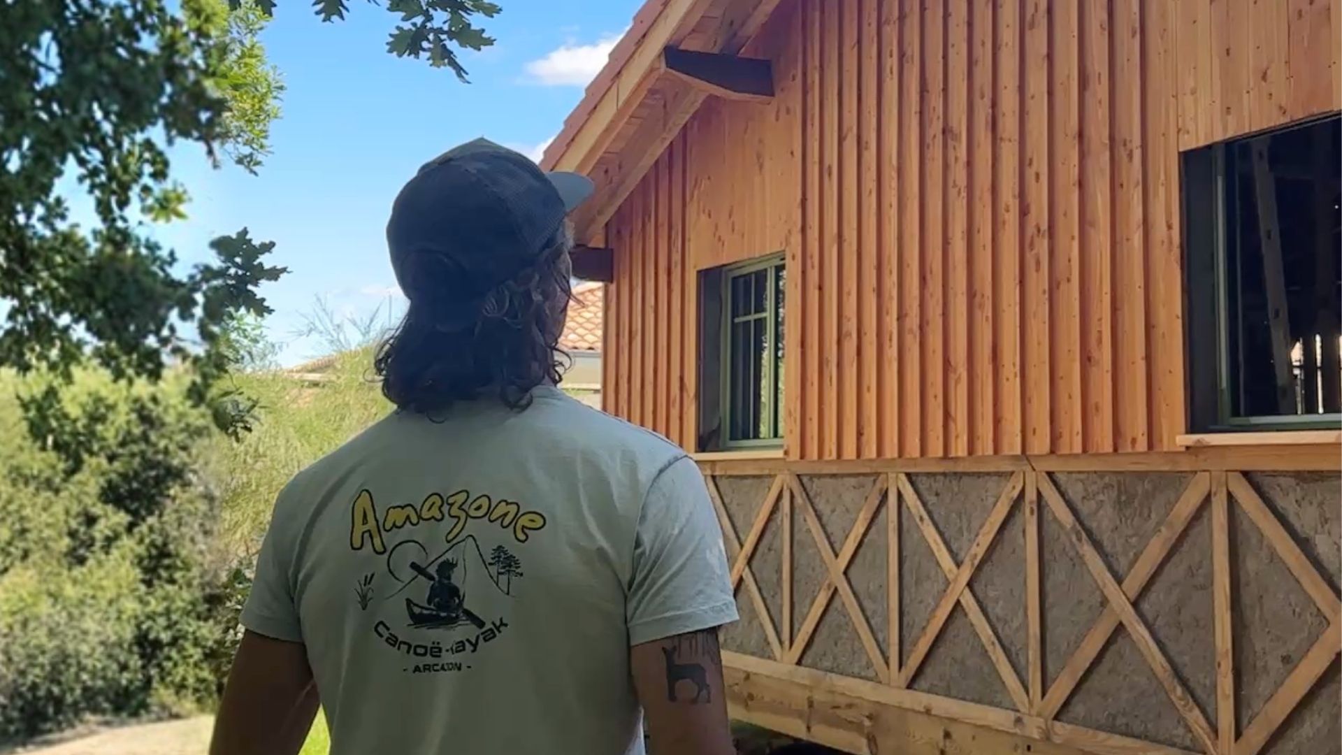 Un homme de dos avec un tee shirt gris et une casquette regarde une maison en bois