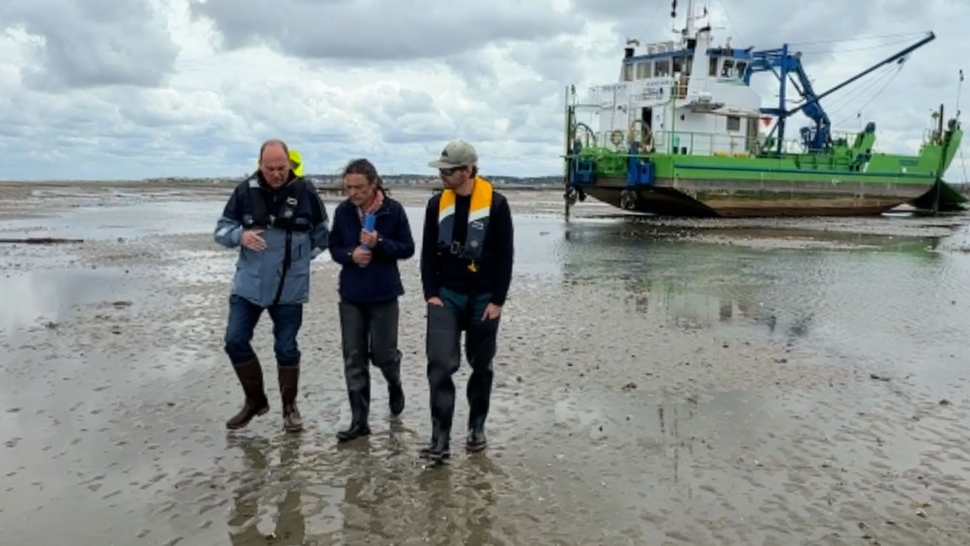 Trois personnes marche sur un banc de sable devant un gros bateau vert. Ciel gris.