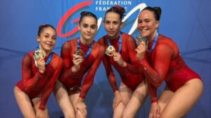Quatre jeunes gymnastes portent des justaucorps rouges assortis et sourient devant un fond bleu avec le logo de la Fédération Française de Gym. Chacune tient fièrement une médaille autour du cou, symbolisant leur réussite dans une compétition de gymnastique.