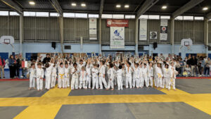 Des enfants sont debout sur un tatami dans un dojo, portent une tenu de judoka et soulèvent des trophées vers le haut.