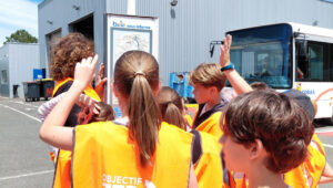 Des enfants avec des gilets oranges devant un panneau d'arret de buc. àL'arrière : un bus