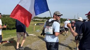 un homme avec un teeshirt bleu et une casquette marche dans l'herbe avec un drapeau francais bleu blanc et rouge. D'autres personnes autour de lui
