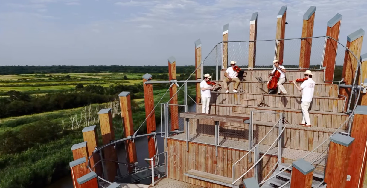 Des musiciens en blanc au sommet d'un belvedaire en bois qui surplombe un paysage de landes vertes à l'occasion du festival des escapades musicales