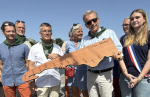 deux hommes tiennent une sculpture en bois représentantla carte de la vile de Lège-Cap ferret pour l'anniversaire d ela commune.Ils sont entourés de nombreuses personnes dont une jeunes femmes portant l'écharpe tricolore et un homme en chemise bleue et pantalon orange