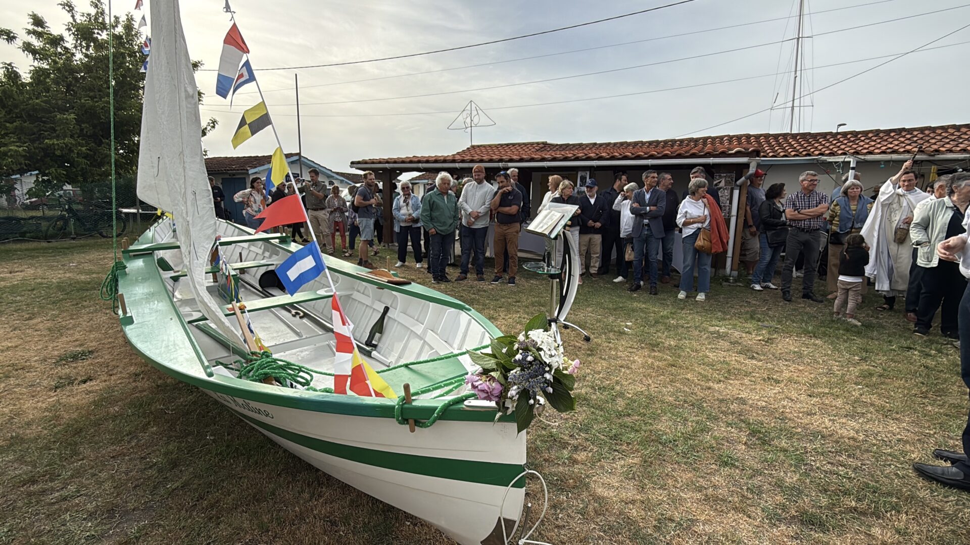 Un bateau en bois blanc et vert avecun mat et une voile posé sur l'herbe devant un groupe de personnes rassemblées