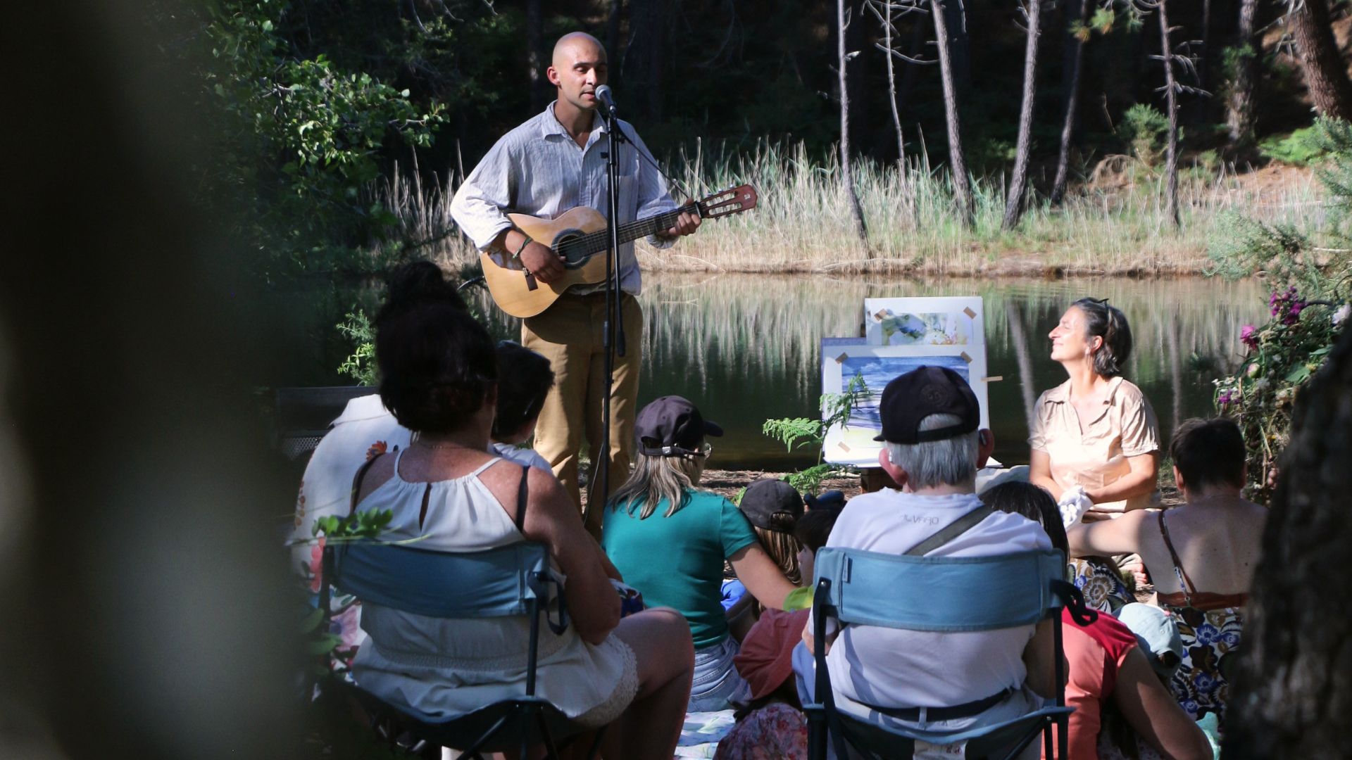 un musicien avec sa guitare dans un espace naturel , un groupe de personne l'écoute entourées d'arbres d'arbustes et d'un plan d'eau