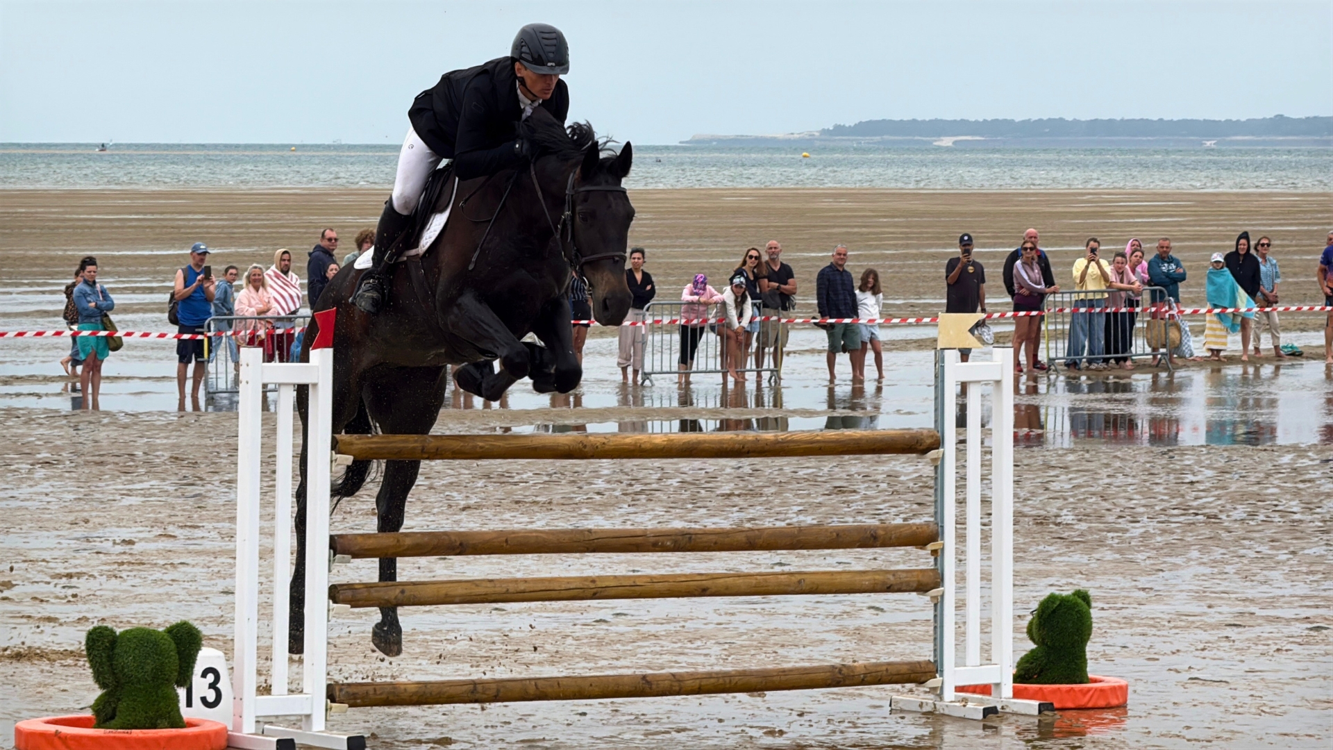 cheval avec son cavalier en train de sauter un obstacle sur la plage avec des spectateurs en arrière plan