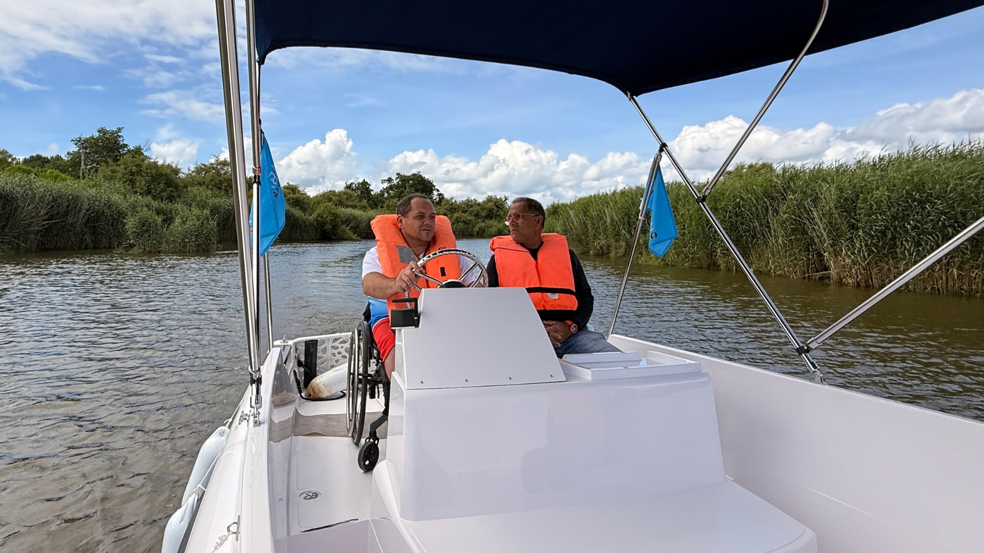 une personne en fauteuil roulant à la barre d'un bateau et un homme assis à côté, en arrière plan, la leyre