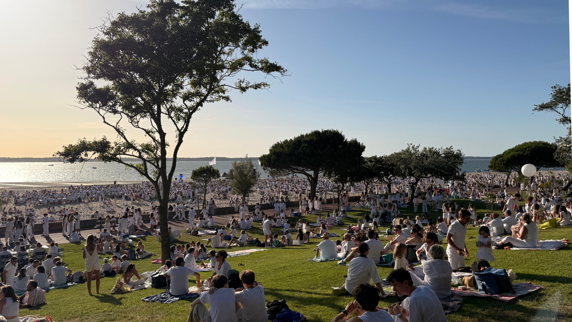 au coucher de soleil, de nombreuses personnes assises sur l'herbe et sur la plage habillées en blanc à l'occasion de la soirée blanche d'arcachon