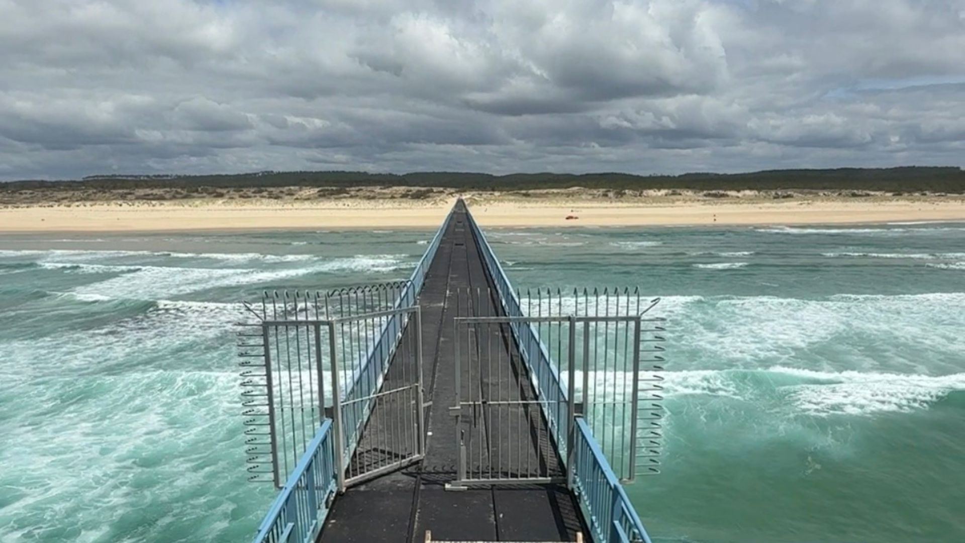 une jetée qui s'avance dans la mer. au loin une plage bordée par la foret