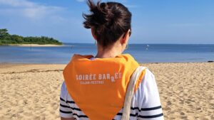 jeune femme brune de dos qui porte une marinière et un bandana orange autour du cou à l'effigie de la soirée barrée de Gujan-Mestras, avec la plage de la hume en arrière plan