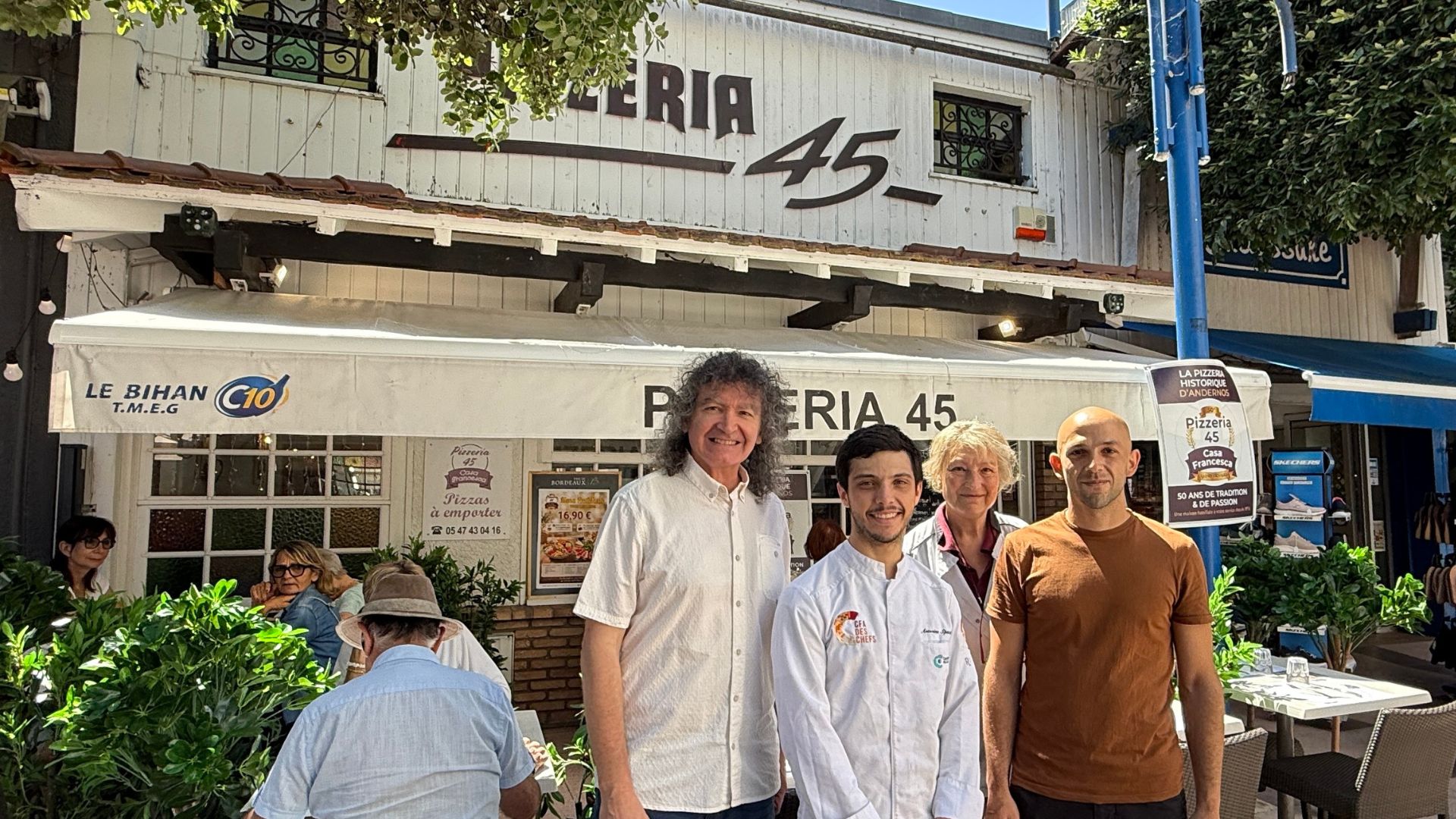 Un groupe de 4 personnes posent devant la terrasse d'un restaurant nommé "Pizzeria 45". Beau temps, des gens en terrasse derrière eux.