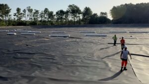 des hommes en tenue de chantier avec des gilets oranges dans un grand bassin vide, sans eau recouvert d'une bâche noire et entouré d'arbres