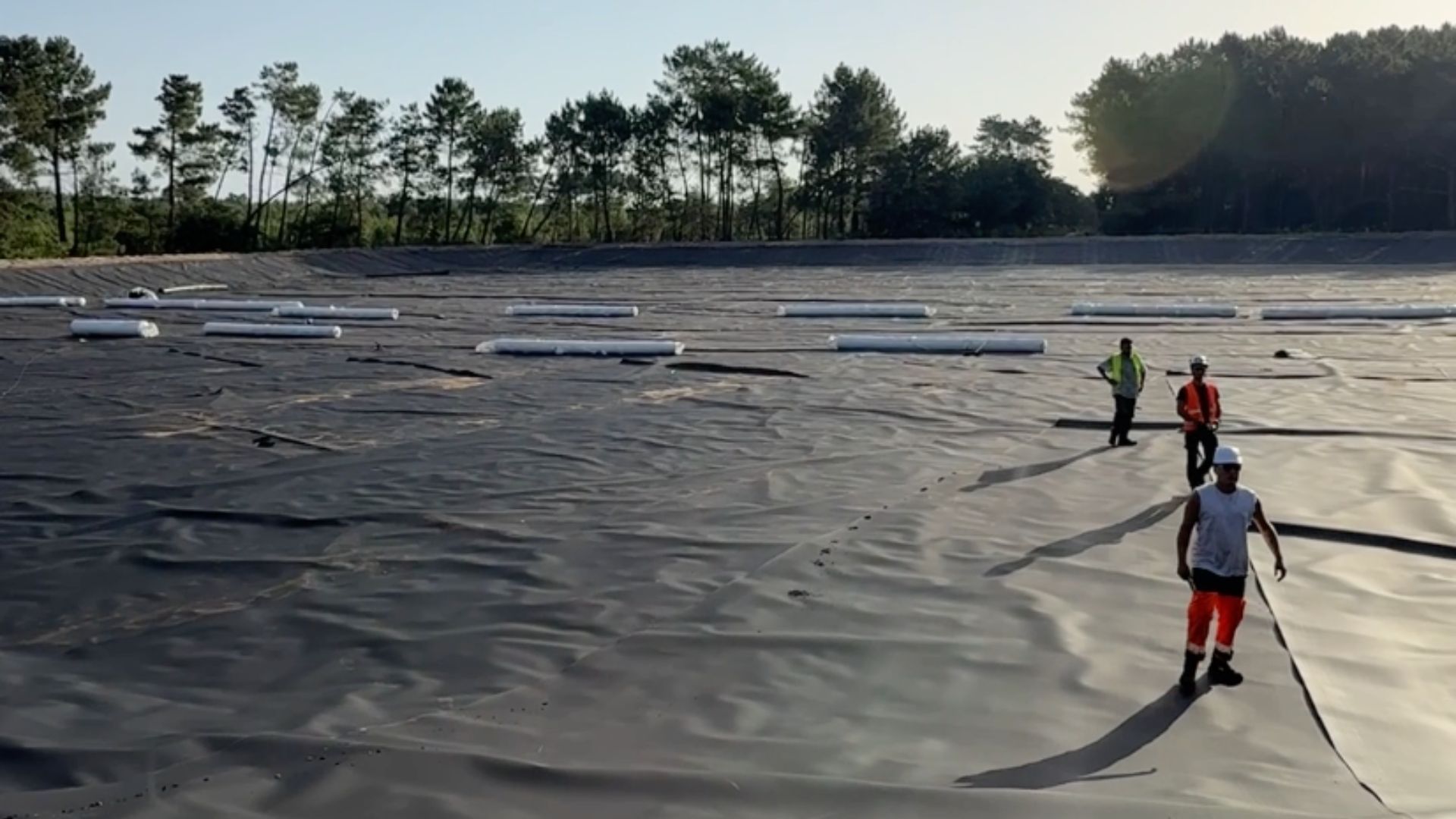 des hommes en tenue de chantier avec des gilets oranges dans un grand bassin vide, sans eau recouvert d'une bâche noire et entouré d'arbres