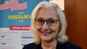 Josette serres : femme souriante avec les cheveux courts et des lunettes. Une affiche en fond