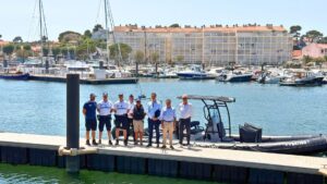des hommes dont certains en unifmre de police sont lignés devant un bateau pneumatique noir. En arrière plan des bateaux amamrées au port et une barre d'immeuble. Image prise en hauteur
