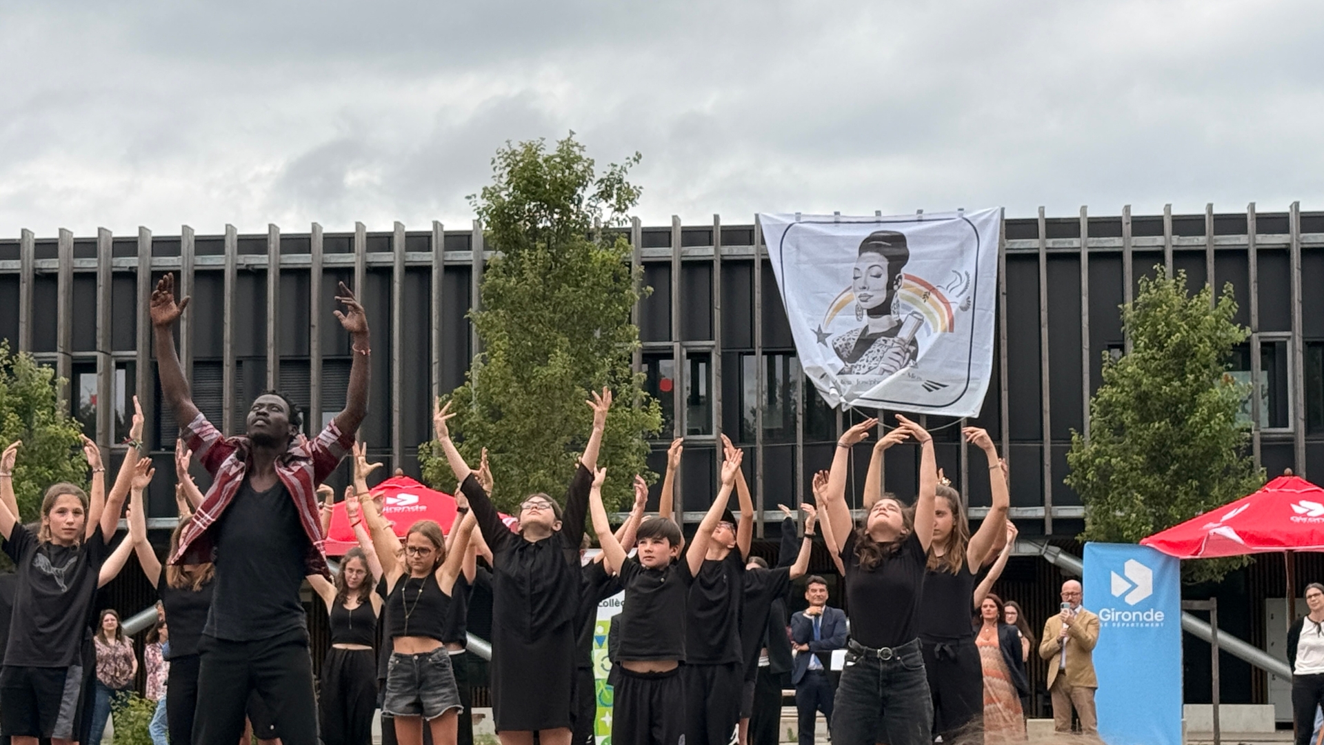 des élèves habillés en noir, les bras vers le ciel. En arrière plan la façade du collège Joséphine Baker de Mios avec le nouveau logo du collège