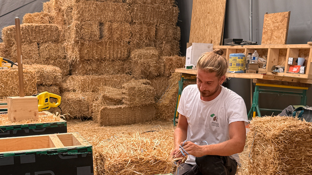 Un homme accroupi manipule de la paille pour la faire entrer dans des ossatures en bois qui servent à la structure d'isolation de la résidence. Derrière lui, plusieurs blocs de pailles sont entreposés.