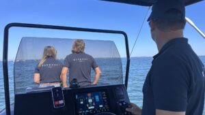 Un homme se tient au poste de pilotage et regarde l'horizon. Derrière le pare-brise un homme et une femme se tiennent côte-à-côte de dos à l'objectif et observent également l'horizon. Sur leurs polos, on voit l'inscription BlueNav.