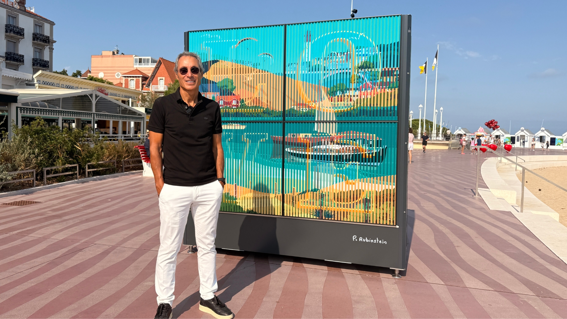 homme avec un polo noir et un pantalon blanc devant une oeuvre artistiques colorée sur le front de mer d'arcachon