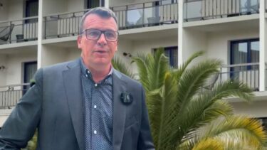 Un homme en costard devant un immeuble avec des balcon. Un palmier derrière lui.
