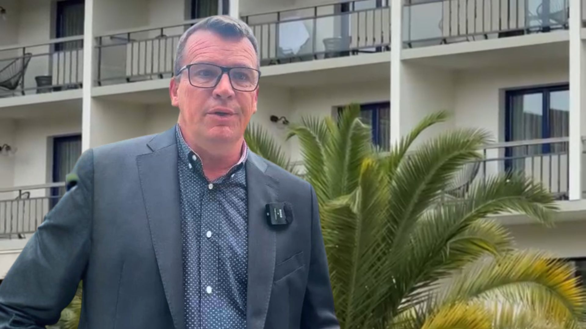 Un homme en costard devant un immeuble avec des balcon. Un palmier derrière lui.