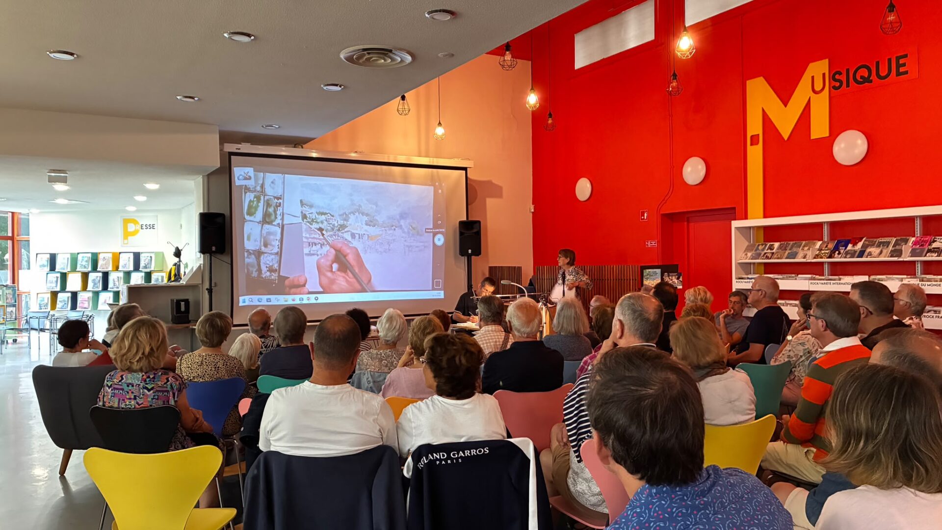 Plusieurs personnes assises assistent à la réalisation d'une aquarelle en direct. Cette animation s'inscrit dans présentation de l'ouvrage d'« Une année sur le Bassin d'Arcachon ».