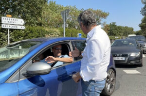 Un homme avec une chemise blanche discute avec un homme dans une voiture bleu. derrière eux des arbustes, ciel bleu