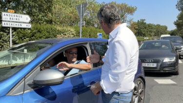Un homme avec une chemise blanche discute avec un homme dans une voiture bleu. derrière eux des arbustes, ciel bleu