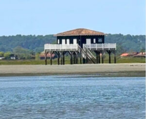 Au premier plan, la mer. Une cabane tchanquée noire et blanche se dresse au second plan de l'image. En arrière plan, un bout de la presqu'île du Cap Ferret et une forêt.