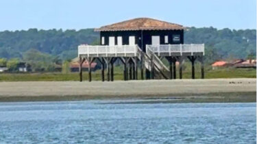 Au premier plan, la mer. Une cabane tchanquée noire et blanche se dresse au second plan de l'image. En arrière plan, un bout de la presqu'île du Cap Ferret et une forêt.