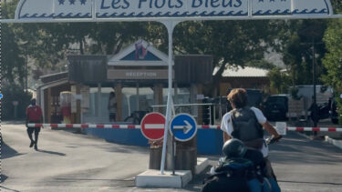 L'enseigne blanche et bleue du camping sur laquelle est inscrite "Camping Les Flots Bleus" prise en photo de face. En arrière-plan, la Dune du Pilat. Un cycliste passe au niveau de la barrière d'entrée.