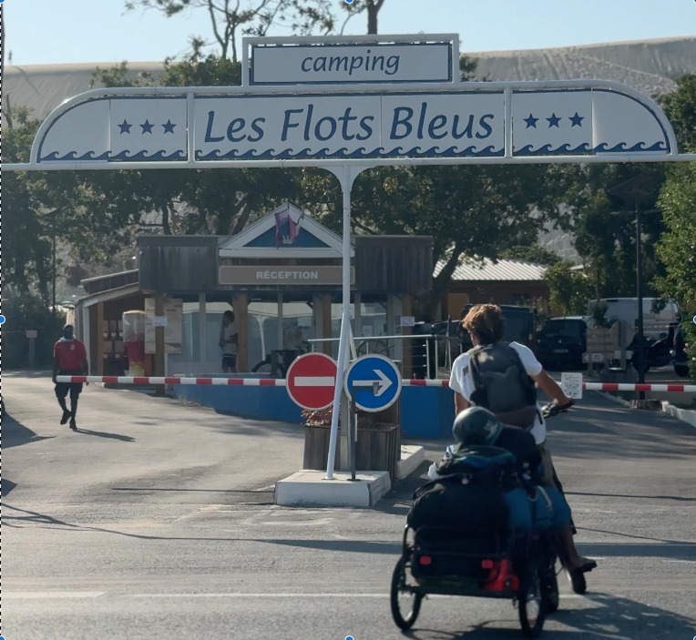 L'enseigne blanche et bleue du camping sur laquelle est inscrite "Camping Les Flots Bleus" prise en photo de face. En arrière-plan, la Dune du Pilat. Un cycliste passe au niveau de la barrière d'entrée.