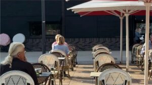 trois personnes à la terrasse d'un café , un mur noir derrière, rayon de soleil et parasols blancs et rouges