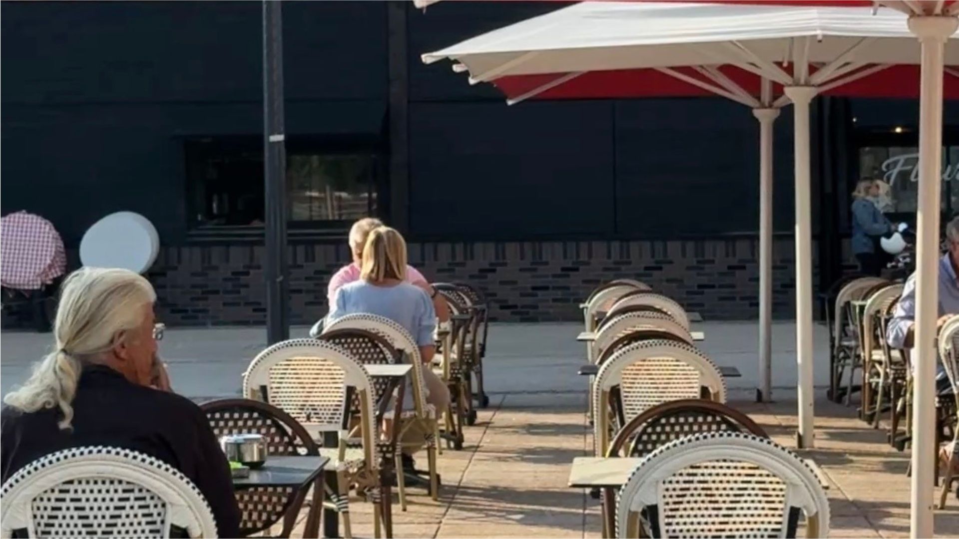 trois personnes à la terrasse d'un café , un mur noir derrière, rayon de soleil et parasols blancs et rouges