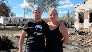 Un homme et une femme se tiennent cote à cote devant une maison brulée, derrière il y a un mobilhome