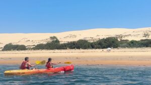 deux personnes dans un canoe rouge et jaune devant une dune de sable